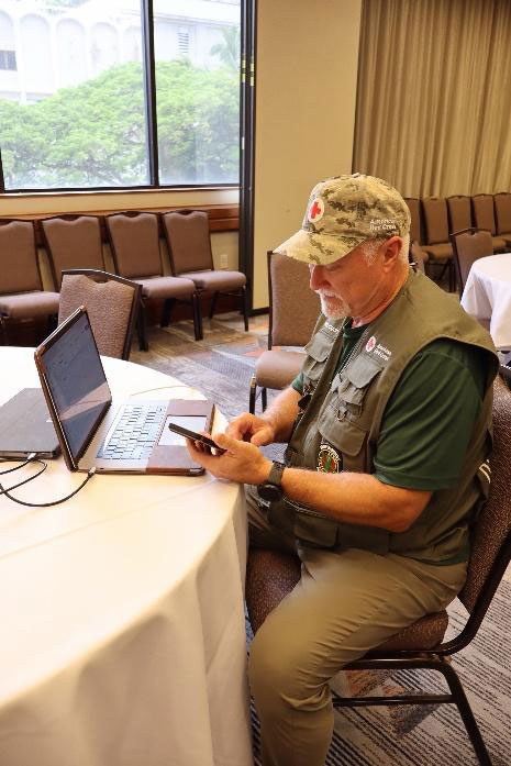Paul Chartier, wearing a Red Cross hat and vest, sitting at a table with a laptop and phone.