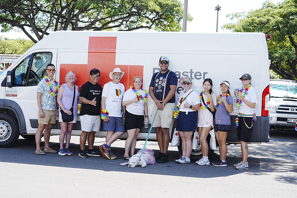 A group of people and a dog at a pride event, wearing pride clothing, standing in front of a Red Cross van.