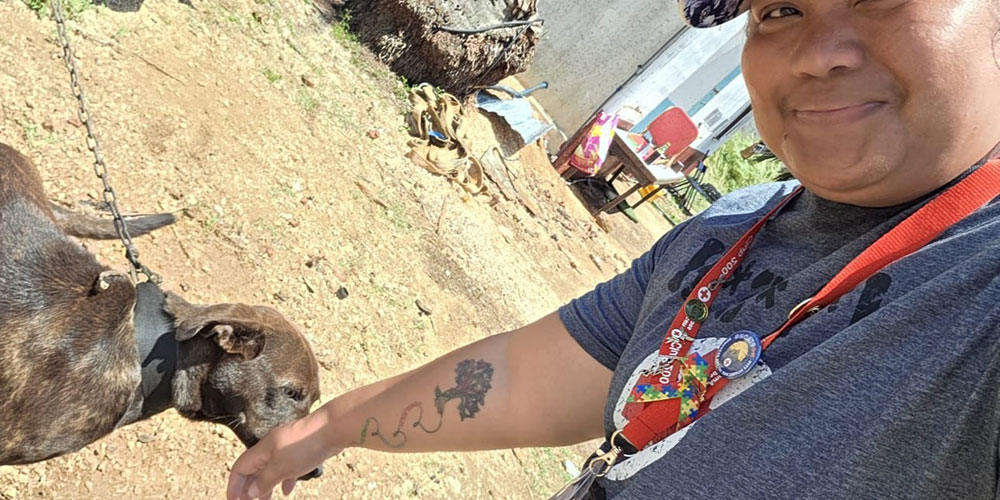 Person wearing a Red Cross shirt and red lanyard with pins and rainbow puzzle ribbons in it reaches toward a brown dog on a leash in a dirt yard.