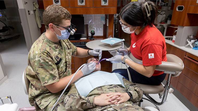 Two dental professionals, one in camouflage and another in a red cross shirt, treat a patient in military uniform in a clinic.