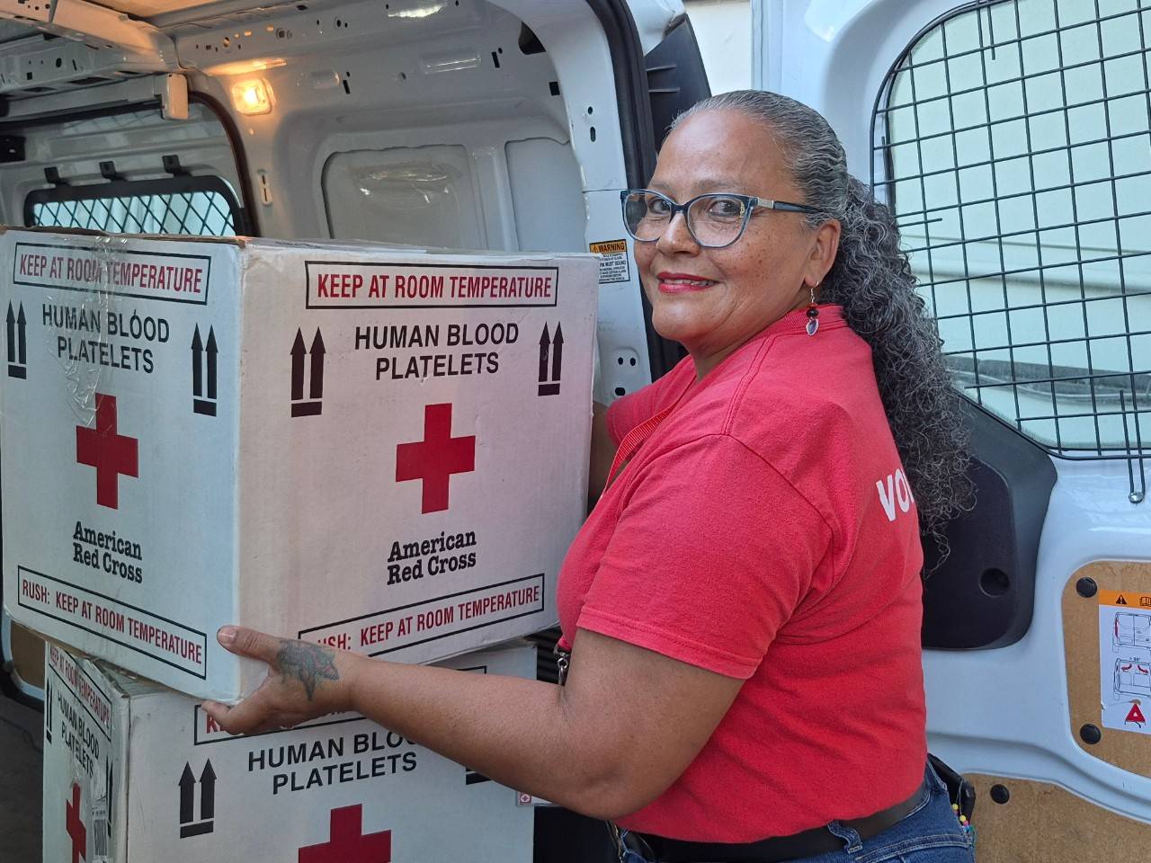 Voluntaria Elizabeth Ríos aguanta una caja de sangre mientras mira a la cámara., frente a un vehículo a punto de cargar la caja.