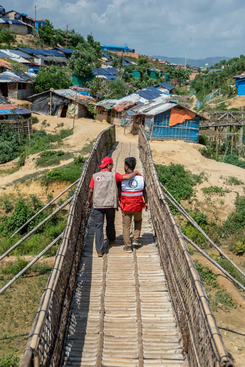 June 29, 2019. Cox s Bazar, Bangladesh. An American Red Cross and Bangladesh Red Crescent team walks across a bamboo bridge in Kutupalong a displacement camp in Cox  Bazar, Bangladesh. Since August 2017, more than 700,000 people have fled Rakhine State, Myanmar to seek safety in Cox s Bazar, Bangladesh. Many arrived injured, malnourished, and devastated. They speak of dangerous journeys walking days on end to reach the border and losing touch with family members along the way. Once in Bangladesh, they crowd into camps on muddy hillsides and live in structures made of bamboo, plastic, cardboard and sometimes corrugated metal sheeting. Monsoon rains and dangerous cyclone seasons put migrants at risk of landslides, floods, and destructive wind. Despite harsh conditions and because there s no possibility of evacuating the displacement camp during cyclones migrants are volunteering to help their neighbors during natural disasters and other emergencies. 

The American Red Cross has been working alongside global Red Cross and Red Crescent teams to provide lifesaving aid to the families. We are training migrants on first aid, early warning systems, and other skills so they can respond to rain, wind, flooding, landslides, and cyclones. As part of the Bangladesh government s Cyclone Preparedness Program (CPP), the American Red Cross, Bangladesh Red Crescent, and IFRC are training thousands of camp residents and local (host) community members. In addition, the Red Cross supports disaster simulation drills and equips volunteers to warn fellow residents of danger via megaphones, a flag system, and even loudspeakers at mosques. Photo by Brad Zerivitz/American Red Cross. 