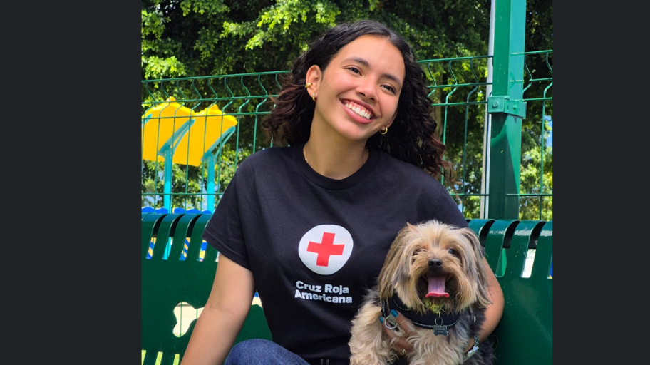 Voluntaria de Cruz Roja sentada en un banco en un parque junto a una mascota en sus brazos.
