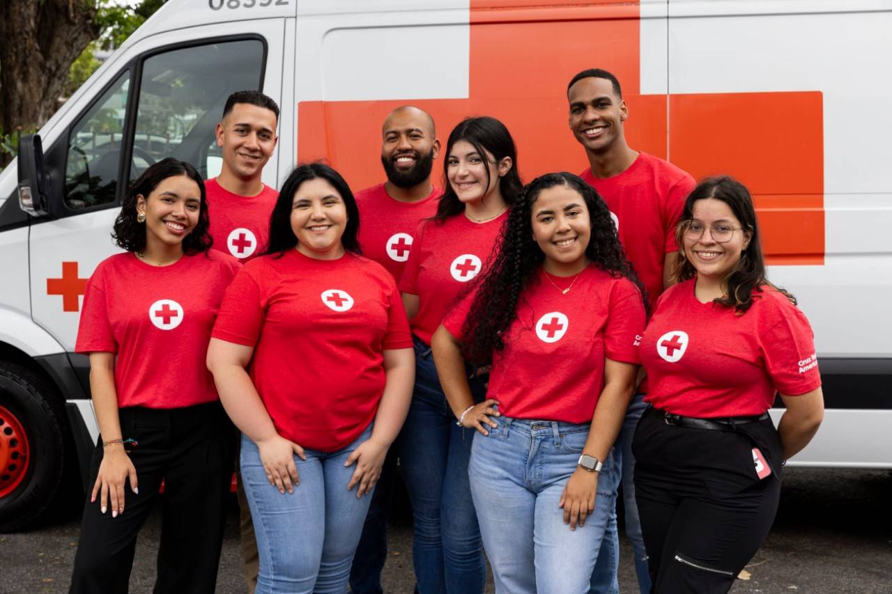 Voluntarios de diferentes edades y géneros sonríen frente a un vehículo de emergencia de la Cruz Roja.
