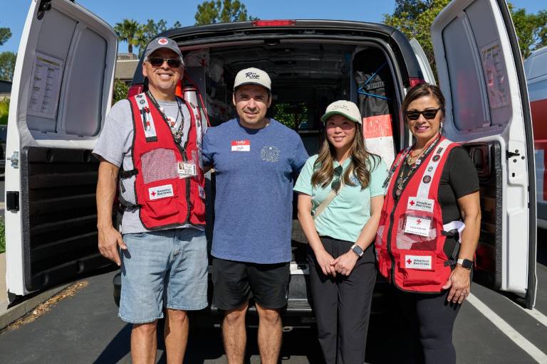 A group of four people stands together in front of an open Red Cross branded response vehicle. Two individuals wear red disaster‑response vests with multiple badges and tags, while another person wears a light-colored shirt and a hat. The fourth person wears a blue shirt and shorts. The interior of the vehicle is visible, showing shelves and supplies. The group is standing in a sunny outdoor parking area with trees and additional vehicles in the background.