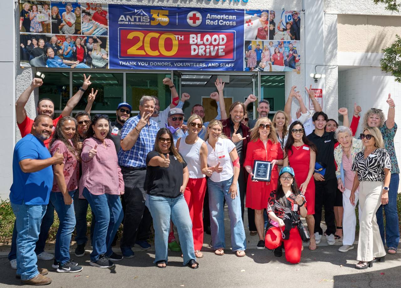 Six adults pose together in front of a blue American Red Cross and Antis Roofing & Waterproofing “35 Years” backdrop, holding blood donation–themed signs. The signs include messages such as “Give Blood Get Cookies,” “I’m the Giving Type,” and “American Red Cross Blood Donor,” with participants wearing red, black, and patterned clothing.