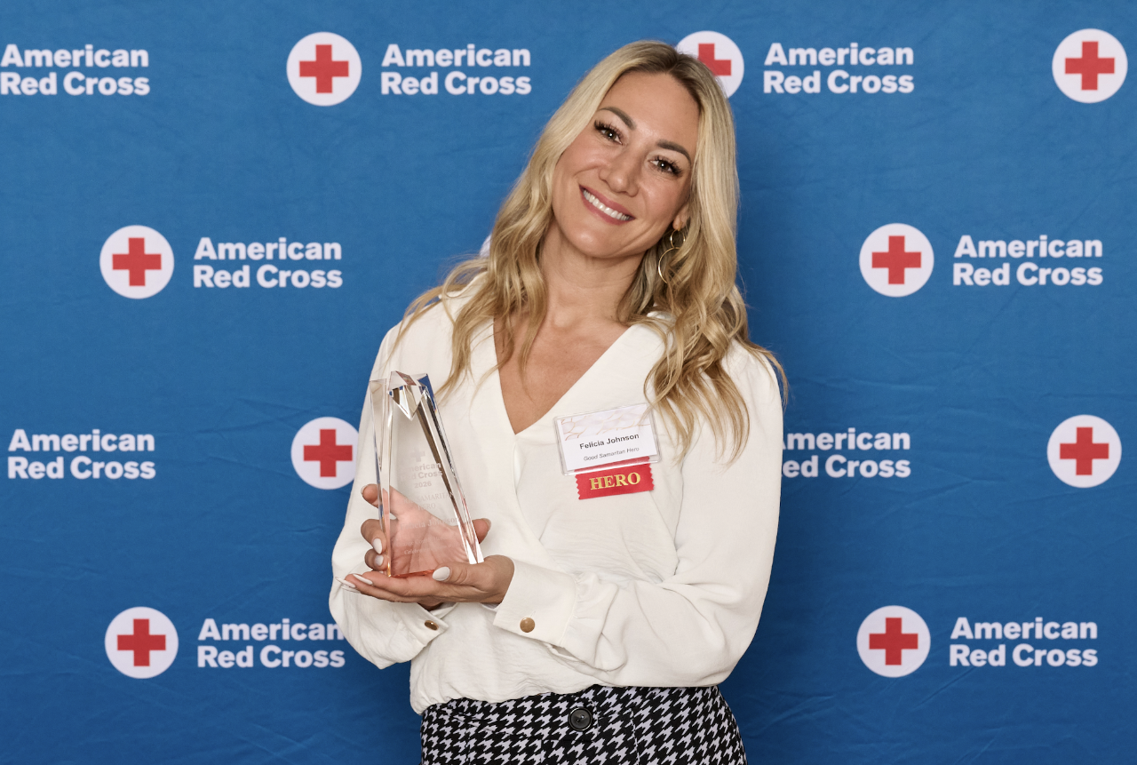 Award recipient holding a glass trophy, standing in front of a blue American Red Cross backdrop with repeating Red Cross logos.