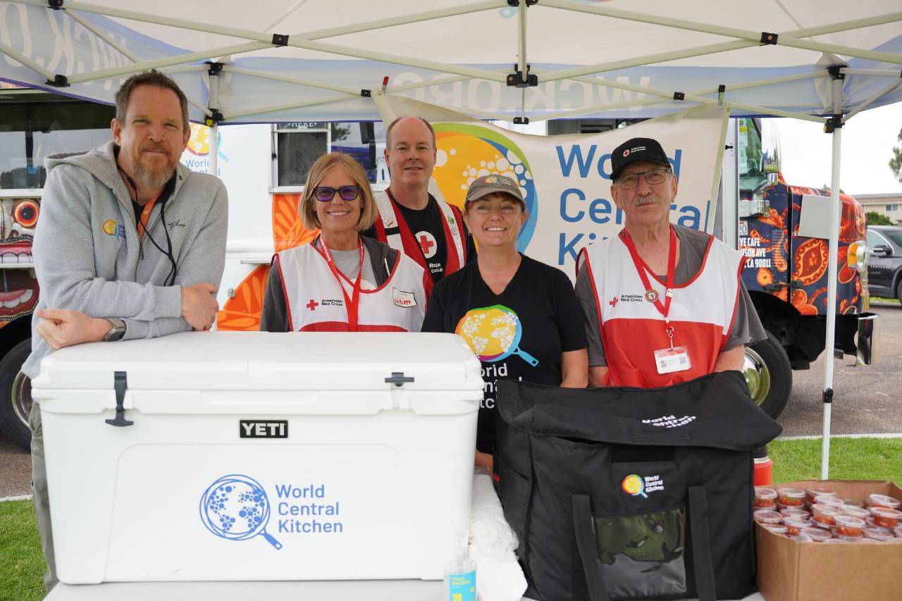 Red Cross volunteers and World Central Kitchen team members stand together with meal supplies and a World Central Kitchen food truck in the background.