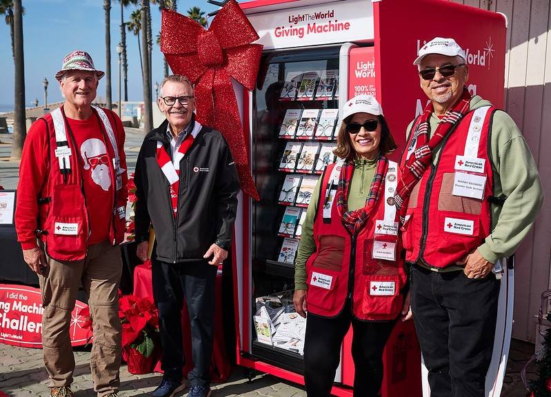 Red Cross volunteers in Red Cross vests standing and smiling in front of the Light the World Giving Machine in Huntington Beach.