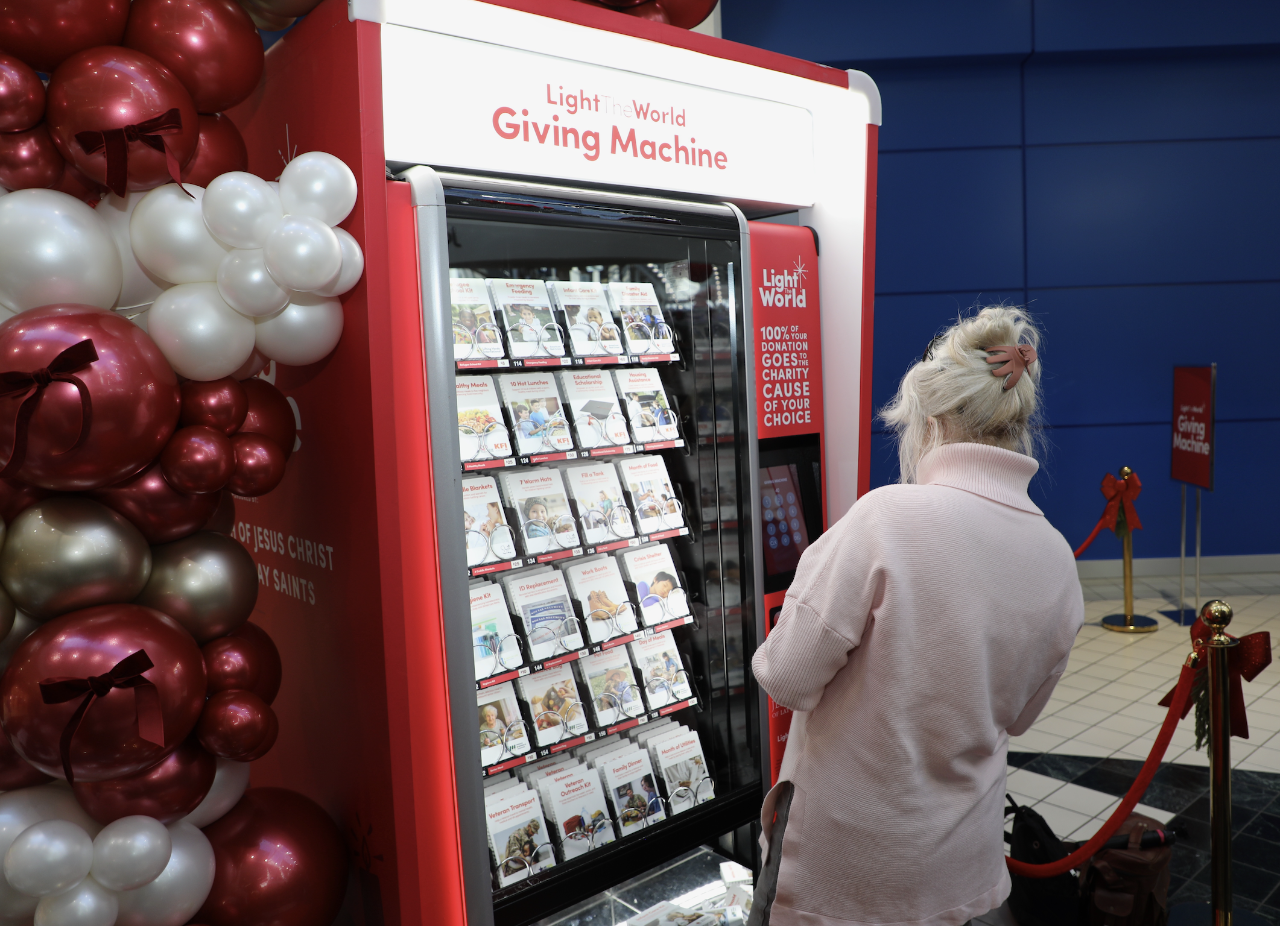 A person stands in front of a red “Light the World Giving Machine” vending-style donation kiosk inside a mall. The machine displays rows of cards showing different charitable donation options. Decorative balloons in red, white, and gold are arranged beside the machine, and stanchions with red ropes surround the area.