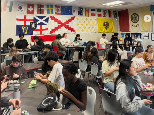A diverse group of students sits at tables in a classroom with international flags on the wall. They are engaged in conversation and eating lunch, creating a lively atmosphere.