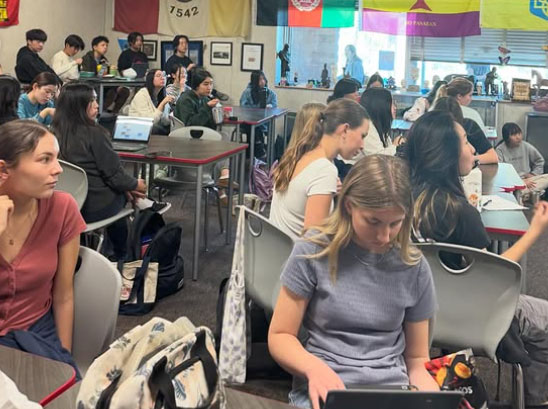 A classroom with students seated at tables, focused on laptops and conversations. Flags and framed photos decorate the back wall.