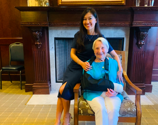 Two women pose for a photo, one sitting in a chair and the other with her arm around her sitting on the arm of the chair
