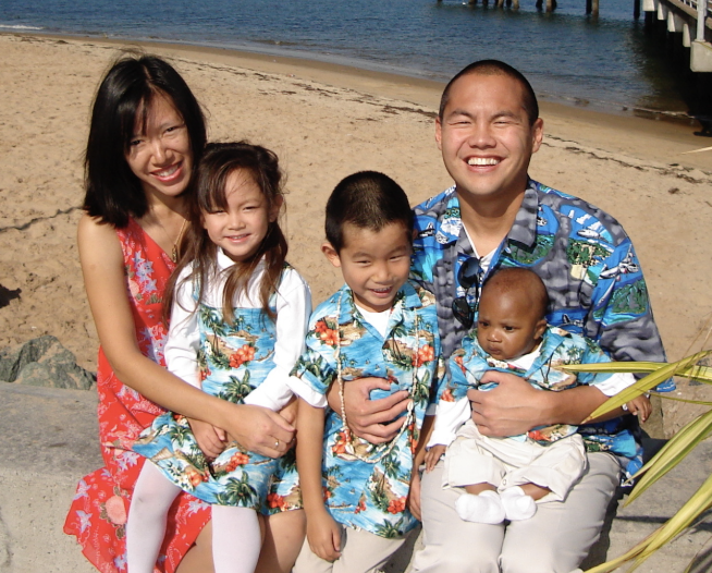 Five people sit together on a concrete edge by a sandy beach with water and a pier in the background. Three smaller children sit between two taller individuals. All five wear colorful, matching or coordinated tropical-print outfits in shades of blue and green.