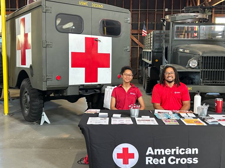 Myka volunteering at a Red Cross table with another Red Cross volunteer. In the background there is an older Red Cross vehicle.