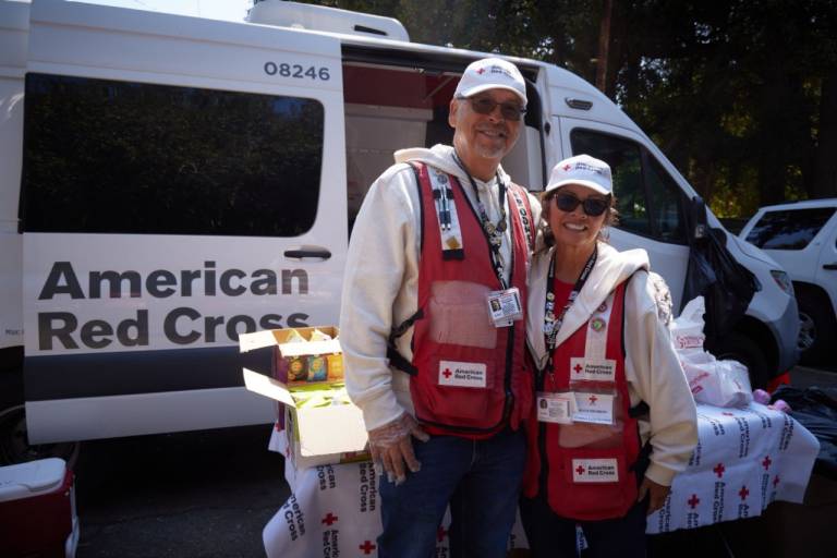 Two American Red Cross workers stand together in front of a Red Cross response vehicle. Both are wearing Red Cross vests, ID badges, light-colored hoodies, and hats. A table beside them is covered with a Red Cross–branded tablecloth and stocked with various supplies, including boxes of snacks. Trees and additional vehicles are visible in the background.