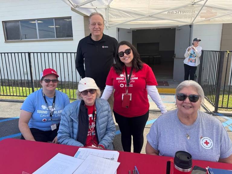 A group of American Red Cross volunteers sit and stand behind a table outdoors under a canopy, with sign-in sheets and materials in front of them.