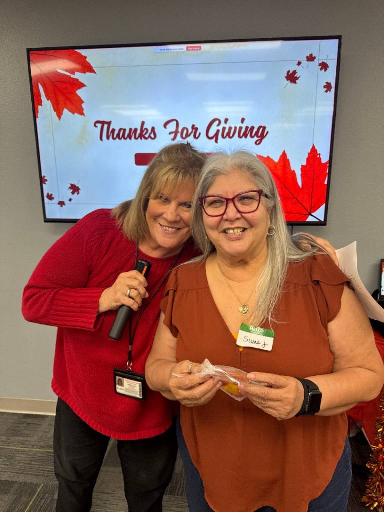 Two American Red Cross volunteers pose together indoors in front of a screen displaying “Thanks For Giving” with fall-themed graphics.