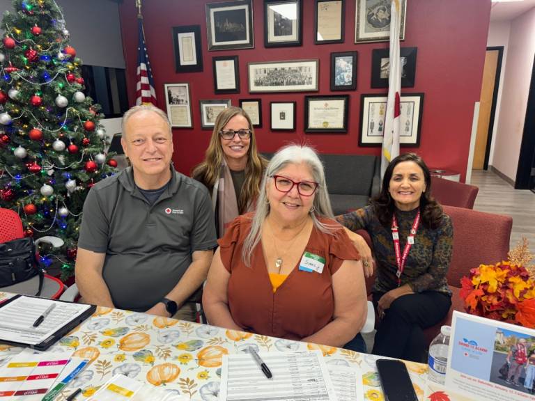 Four American Red Cross volunteers gather around a table indoors near a decorated holiday tree, with forms, pens, and event materials spread out in front of them.
