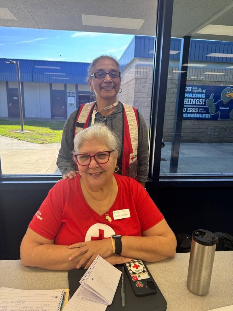 Two American Red Cross volunteers are at a table inside a building near a window, with notebooks, a phone, and a travel mug visible.