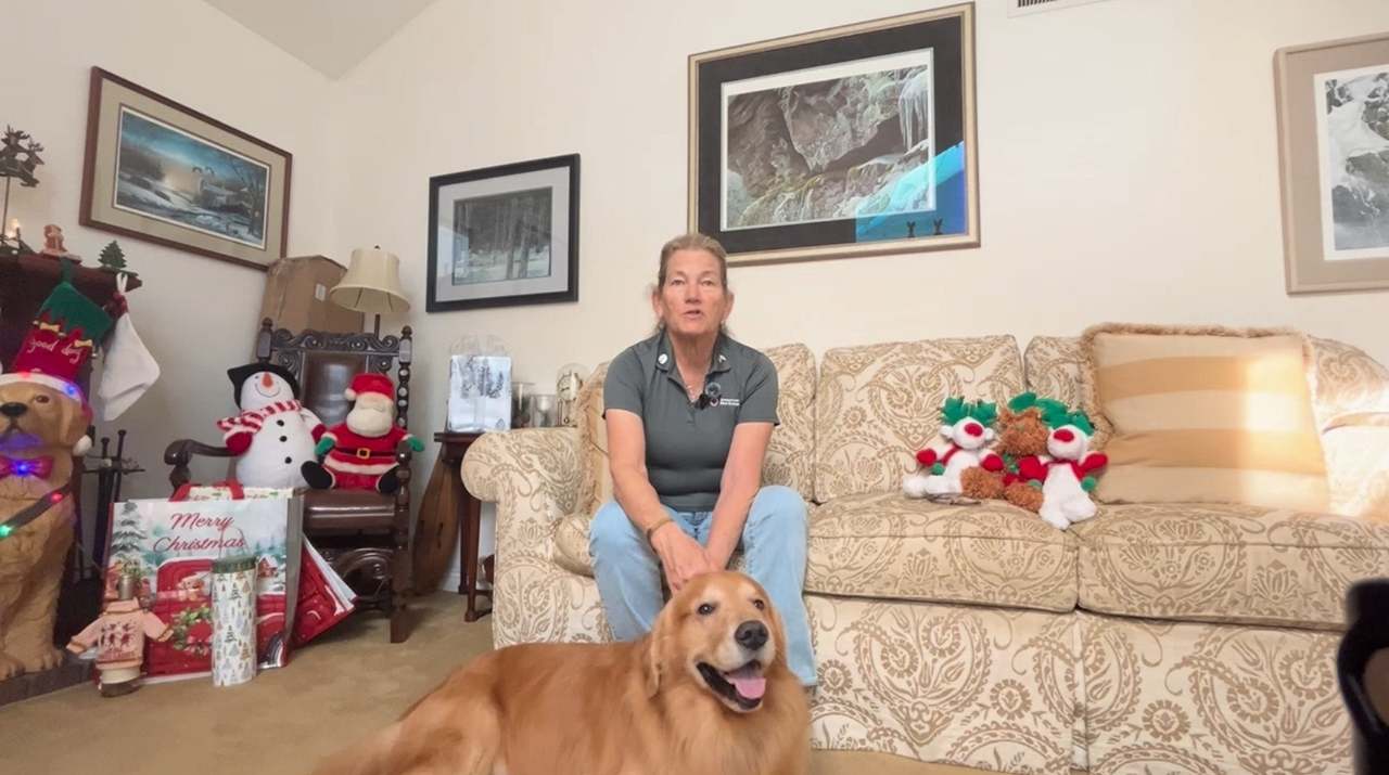 Jan who is a Red Cross volunteer sits on her couch with her golden retriever Teton below her