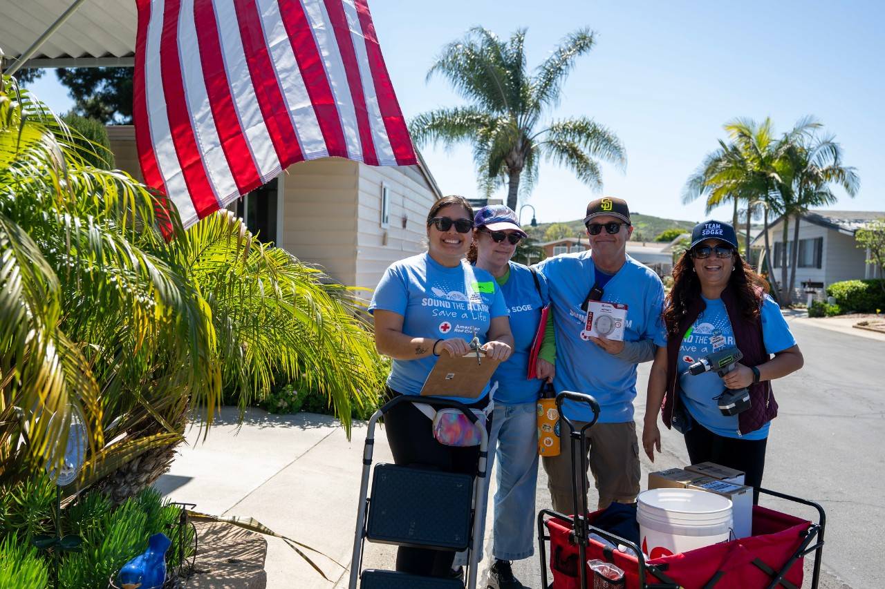 Four American Red Cross volunteers stand on a sunny residential street next to a large U.S. flag and palm trees, wearing blue “Sound the Alarm: Save a Life” shirts and holding tools and supplies, including a clipboard, a smoke alarm, and a drill, alongside a wagon filled with equipment.