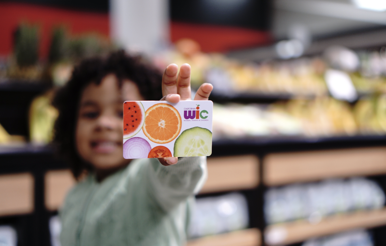 Child in the grocery story produce section holding up a small card that says WIC on the front