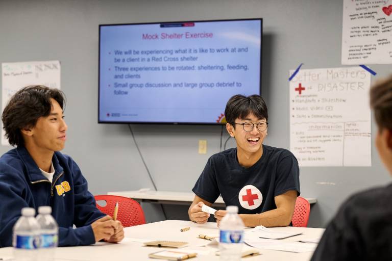 Two youth Red Cross volunteers collaborate in a classroom. One individual is in focus wearing a Red Cross t-shirt and smiling.