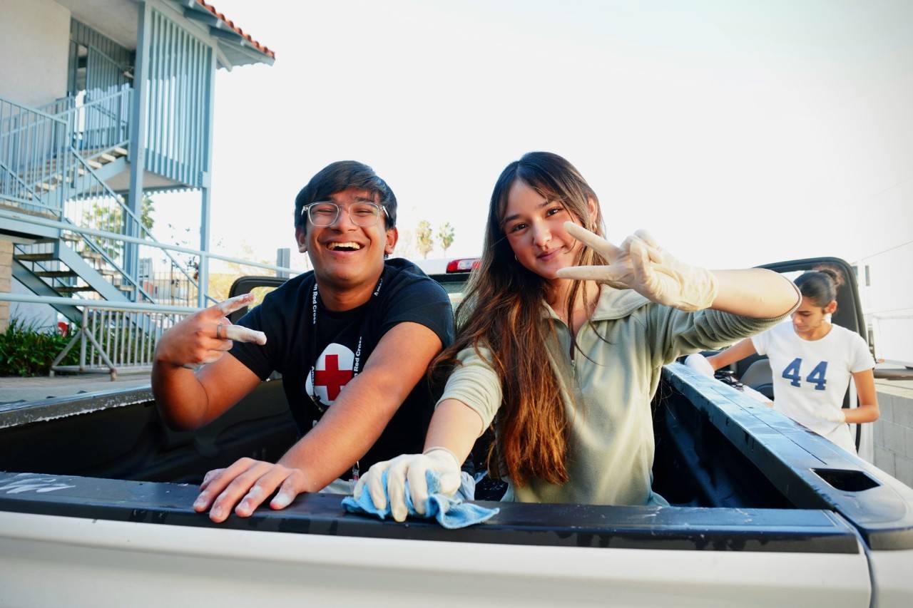 Two Red Cross youth volunteers smiling and giving peace signs while cleaning a Red Cross car