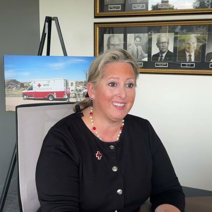 Ellen Vaughan sitting at a table, wearing a Red Cross pin, with a picture of a Red Cross Emergency Response Vehicle behind her.