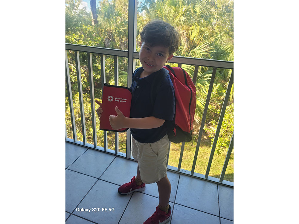 Yound boy wearing a backpack and holding a Red Cross pencil and pen case, giving a thumbs up gesture.