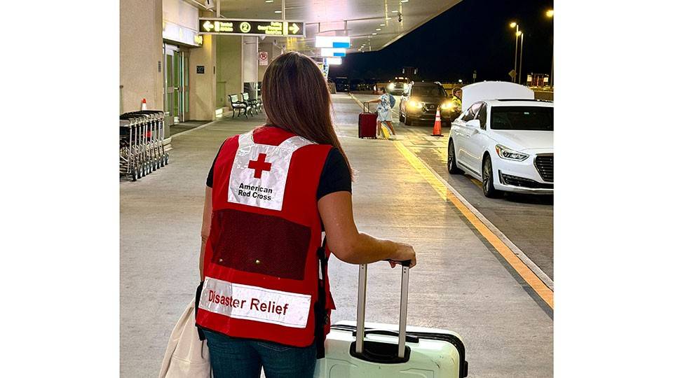 a Red Cross volunteer at the entrance of an airport at night with luggage in hand.