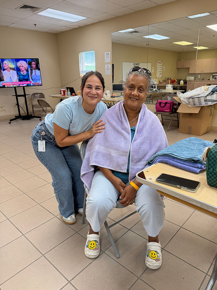 Mercedes Urbina sitting in a chair in a room, with a towel drapped around her shoulders, and another woman leaned over next to her. Both are smiling.