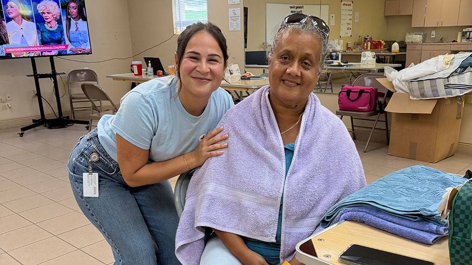 Mercedes Urbina sitting in a chair in a room, with a towel drapped around her shoulders, and another woman leaned over next to her. Both are smiling.