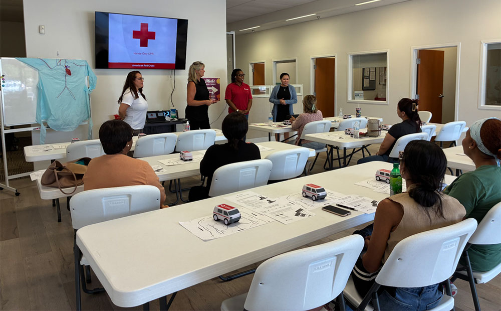 a room with people sitting in chairs listening to Red Cross speakers. A TV on the wall displays the Red Cross logo.