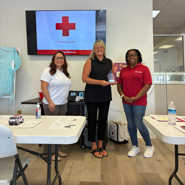 three Red Cross members in a room with one holding an award. A TV on the wall displays the Red Cross logo.