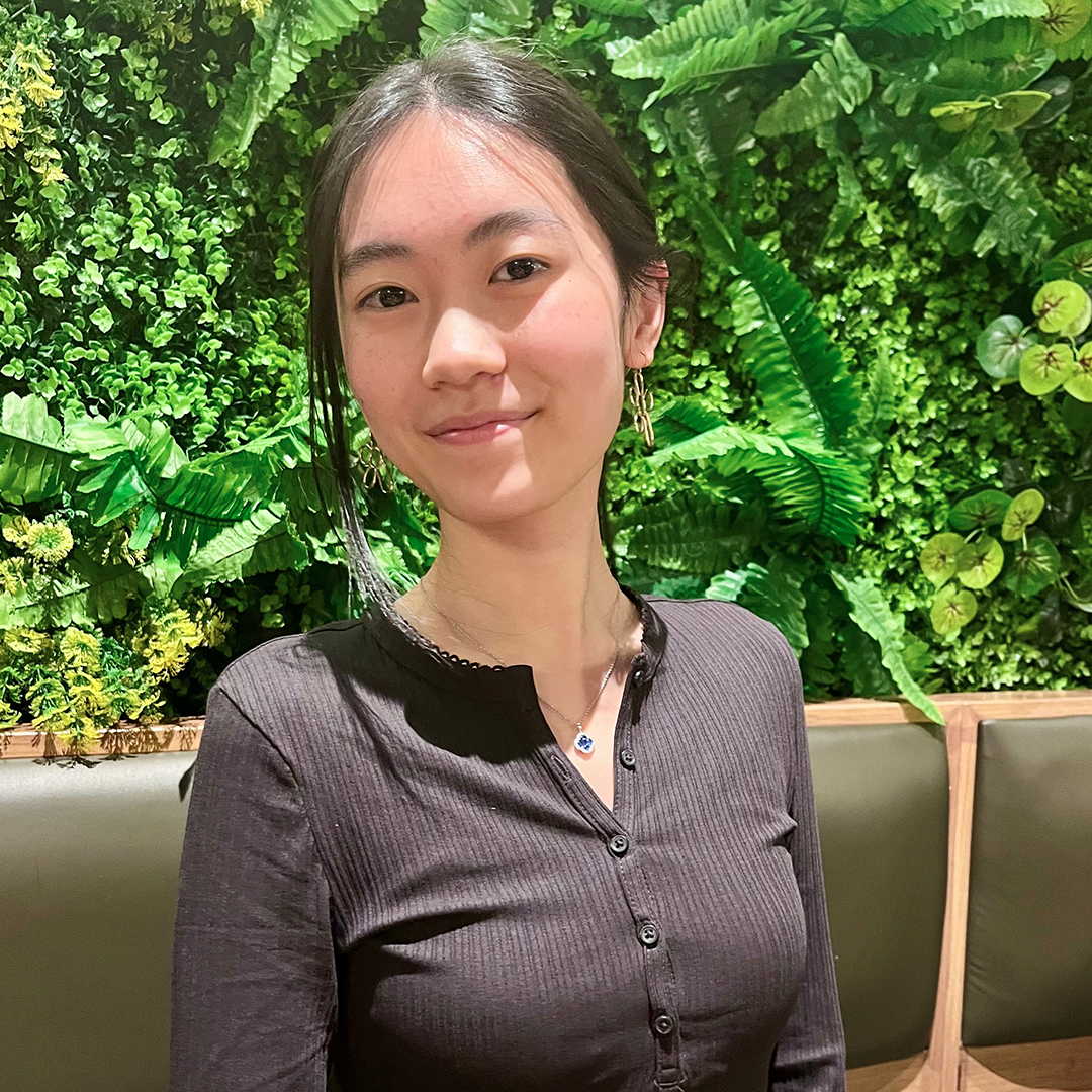 Annie Zhong smiling indoors in front of a green plant wall, wearing a long-sleeved dark grey ribbed top.