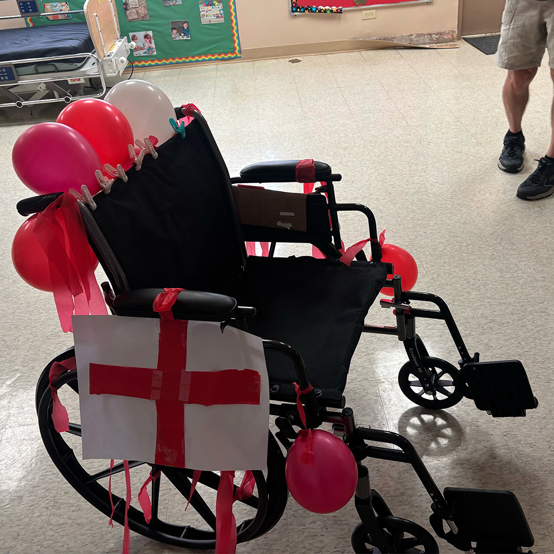 A black wheelchair decorated with red and white balloons and a red cross emblem taped to the front, sitting indoors with people standing nearby.