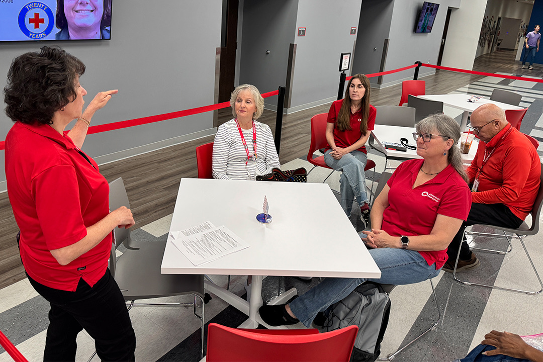 Inside a modern office space, five Red Cross volunteers gather around a small white table for a briefing. On the left, Assistant Director of Operations Debbie Tevlin stands wearing a red shirt and black pants, gesturing with her hand as she speaks. Seated around the table are regional mass care manager Heather Gale, deputy job director Marty Westermann, and ambassadors Kim Majczan and Bonnie Baker. In the background, a flat-screen TV mounted on the wall displays a commemorative "20 Years of Service" graphic.