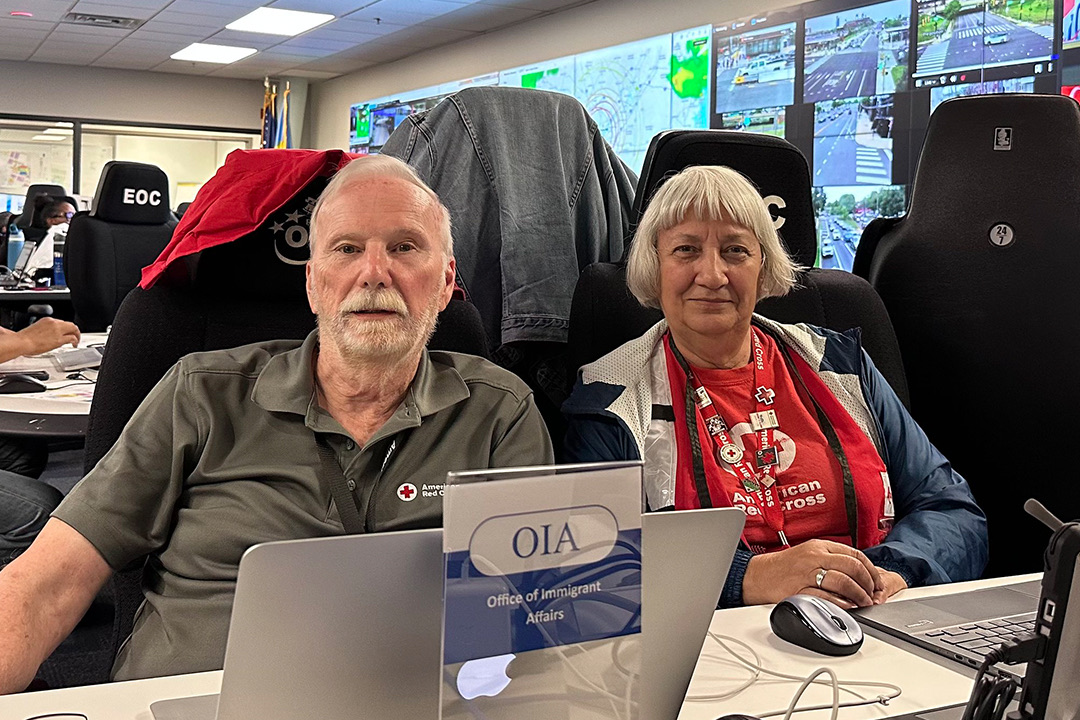 Two smiling Red Cross volunteers sit side-by-side at a desk indoors. On the left, Bruce Swift wears a grey polo shirt with a Red Cross logo, and on the right, Sandy Capoferri wears a red Red Cross lanyard over a white zip-up sweater. A laptop and a small desk sign reading "OIA Office of Immigrant Affairs" sit in front of them. In the background, a large wall is filled with multiple massive television screens showing traffic and surveillance feeds, with an "EOC" sign visible on a desk to the left.