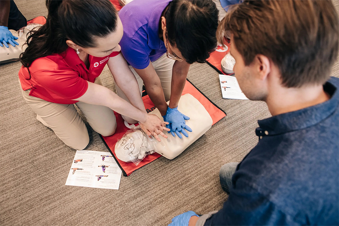 Red Cross instructor in a red shirt guiding a participant through CPR hand placement on a practice mannequin during a training class.