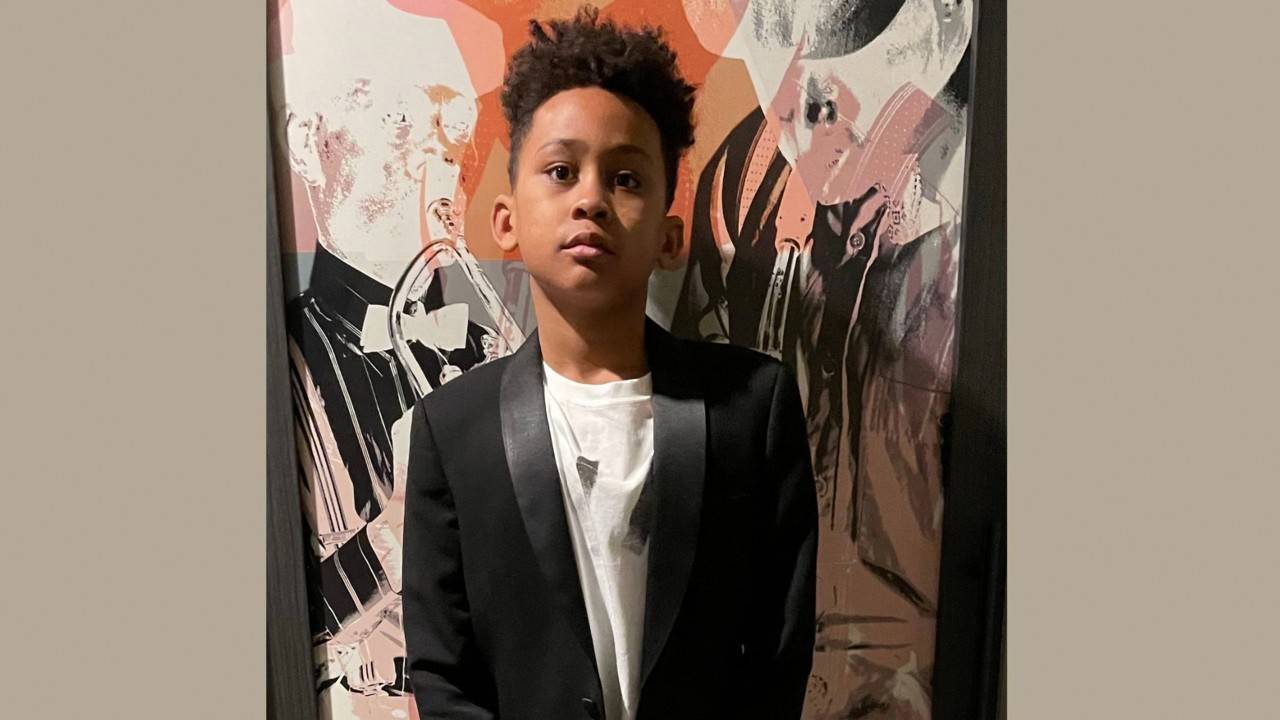 Head to toe photo of 14-year-old Cameron Williams in front of an abstract painting. He is wearing a black coat and pants, white T-shirt and black and white sneakers.