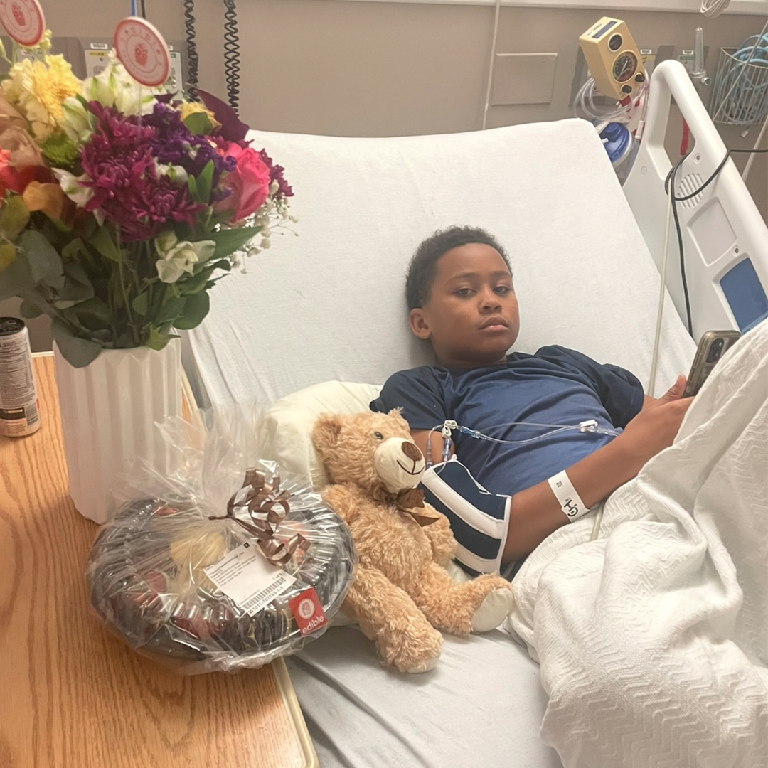 Photo of a young Cameron Williams in a hospital bed with a stuffed bear beside him. He is wearing a blue short and is half covered with a white blanket. A vase of flowers and another wrapped gift sit beside him on a table.