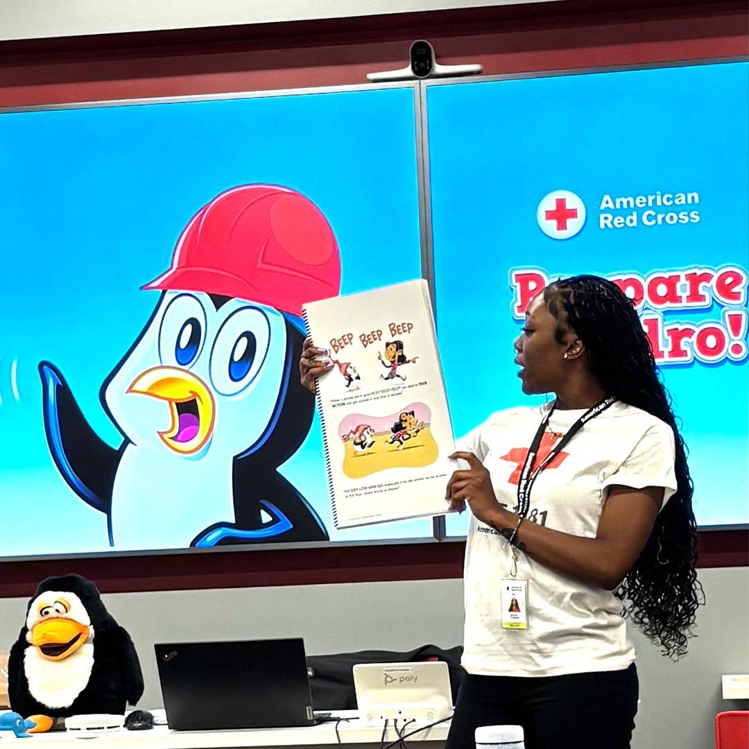 Young Black woman teaches Prepare with Pedro holding up an illustrated book and standing in front of a colorful cartoon penguin graphic.