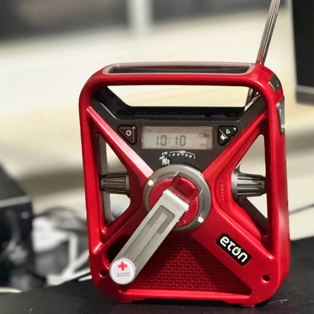 Red and black Etón solar and hand-crank emergency radio with a fold-out hand crank, digital display, antenna, and American Red Cross logo.
