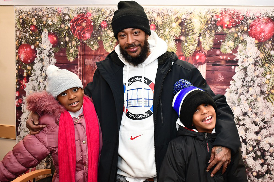 An African American man stands with two elementary school aged children in front of a Christmas background with holly, trees and all ornaments. The three are wearing winter coats and hats. 