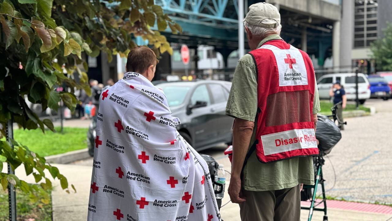 A man in an American Red Cross Disaster Services vest stands with a woman wrapped in a Red Cross blanket in an ourdoor urban environment.
