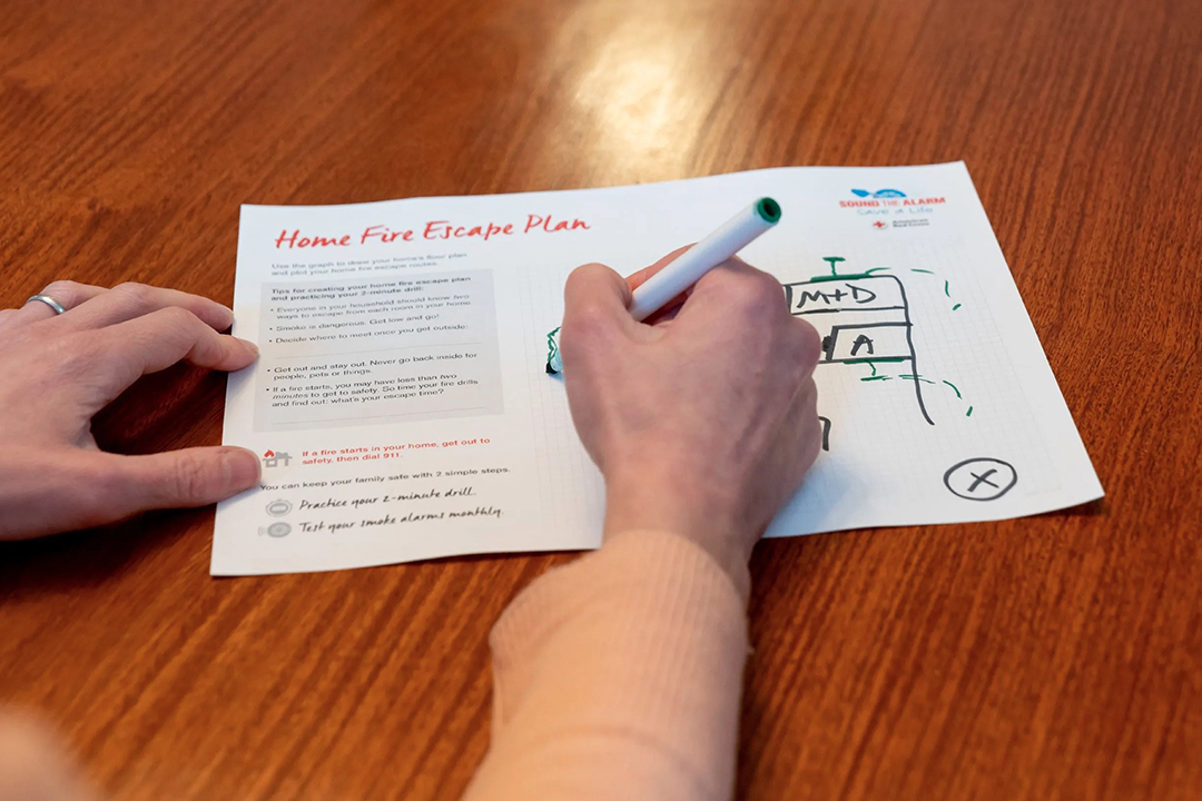 Close-up of a person drawing a home fire escape plan grid next to a printed Red Cross guide on a wooden table.