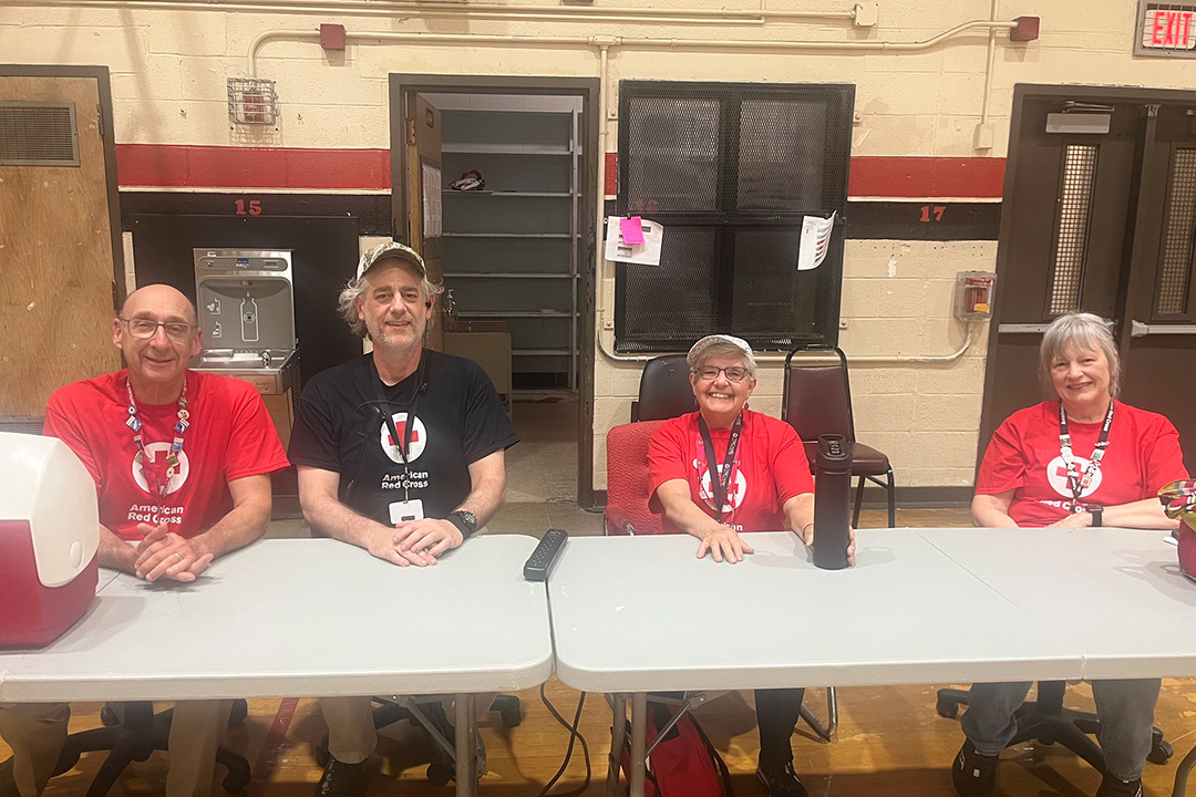 Four smiling Red Cross volunteers sit behind a long white folding table indoors. From left to right, a man in a red Red Cross T-shirt sits next to a cooler, a man in a black Red Cross T-shirt wearing a baseball cap sits with his hands clasped, a woman in a red Red Cross T-shirt rests her hands on the table, and a woman in a red Red Cross T-shirt sits on the far right. Closed lockers and double doors are visible in the background.