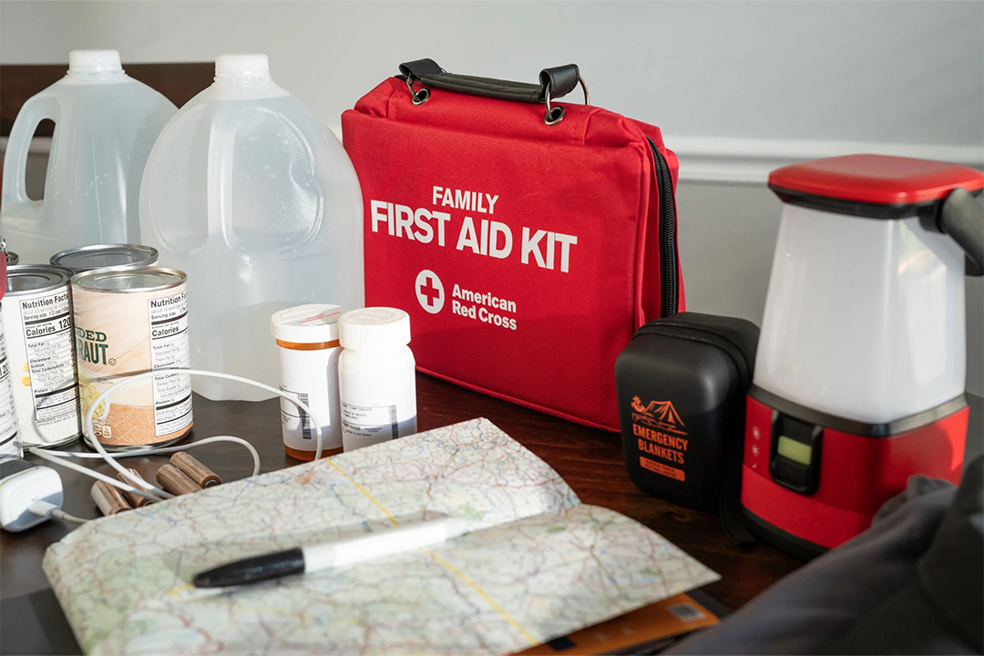 An emergency kit set up on a table featuring water jugs, canned goods, a red American Red Cross family first aid kit, prescription bottles, a lantern flashlight, and an emergency blanket.
